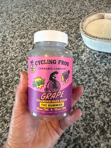 Reviewer's hand holding a bottle of Cycling Frog Super Sour Grape x Hawaiian Haze Rosin THC Gummies.
