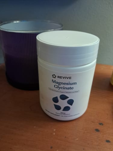 Customer's white bottle of Revive Magnesium Glycinate supplement sitting on a wooden surface next to a dark cup.