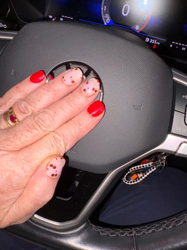 Reviewer's hand showing Cherry Bomb gel nail strips with red and spotted patterns while resting on a steering wheel.