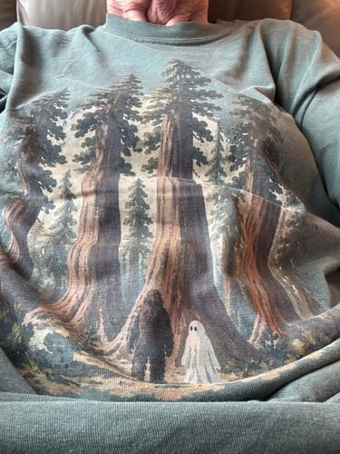 Customer's green Woods We Trust T-Shirt featuring a forest scene with a Sasquatch and a ghost, worn while sitting.