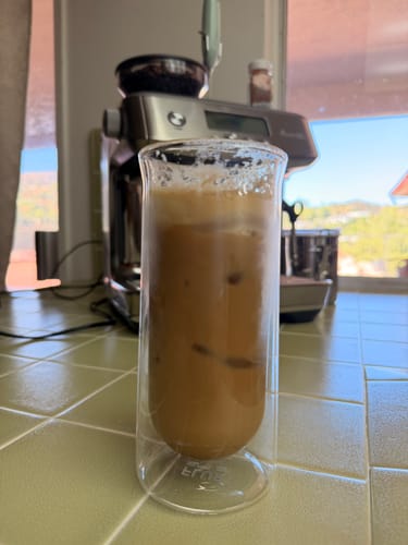 Customer's iced coffee in a clear double-walled FLUR glass, placed on a tiled kitchen counter in front of a coffee machine.