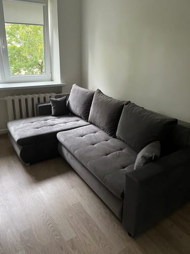 Kliento tamsiai pilka kampinė sofa „Baldo“ pastatyta kambaryje prie lango.