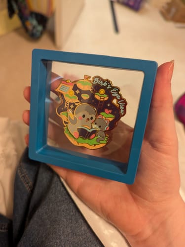 Customer's Birb's Eye View Pin held in a blue square display frame showing a character reading a book.