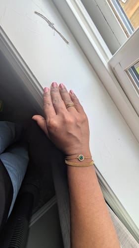 Customer's hand wearing the Roman 18K Gold Bangle and an additional gold-toned bracelet with a green gemstone.