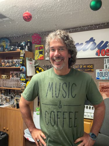Customer's green Coffee and Music T-Shirt worn by a smiling person in a room with shelves of collectibles.