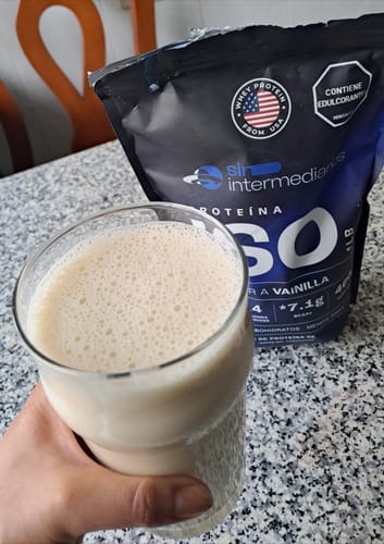 Reviewer's hand holding a glass of creamy vanilla Proteína Aislada next to the protein powder pouch.
