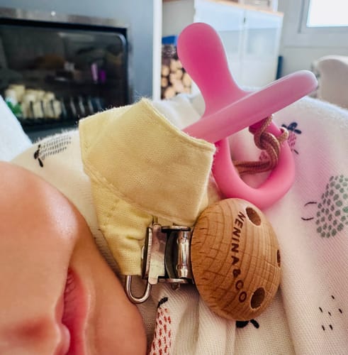 Customer's pink Nenina & Co Chupete Clásico attached to a wooden clip and fabric strap on a baby blanket.