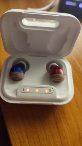 Reviewer's pair of blue and red hearing aid devices inside an open white charging case with glowing status lights.