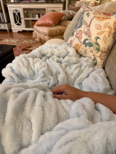 Customer's Hugs Light Shaved Ice Blanket, a soft, pale blue throw draped over their lap while relaxing on a couch.