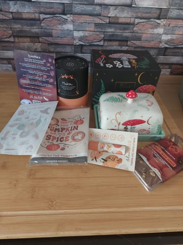 Reviewer's Witchy Box containing mushroom-themed butter dish, ritual candle, lip balm, and autumnal treats on a table.