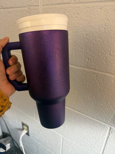 Customer's hand holding a purple glitz Thicc Buddy mug with a white lid against a brick wall.